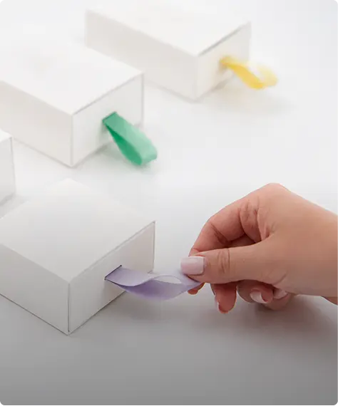 Main saisissant une languette de ruban mauve intégrée à un coffret blanc — Maison Satab