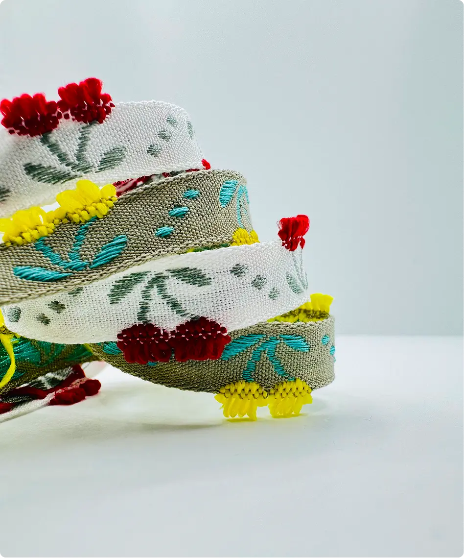 Galons jacquard à motifs floraux verts et turquoise, bordés de franges rouges et jaunes, enroulés sur fond clair — Maison Satab